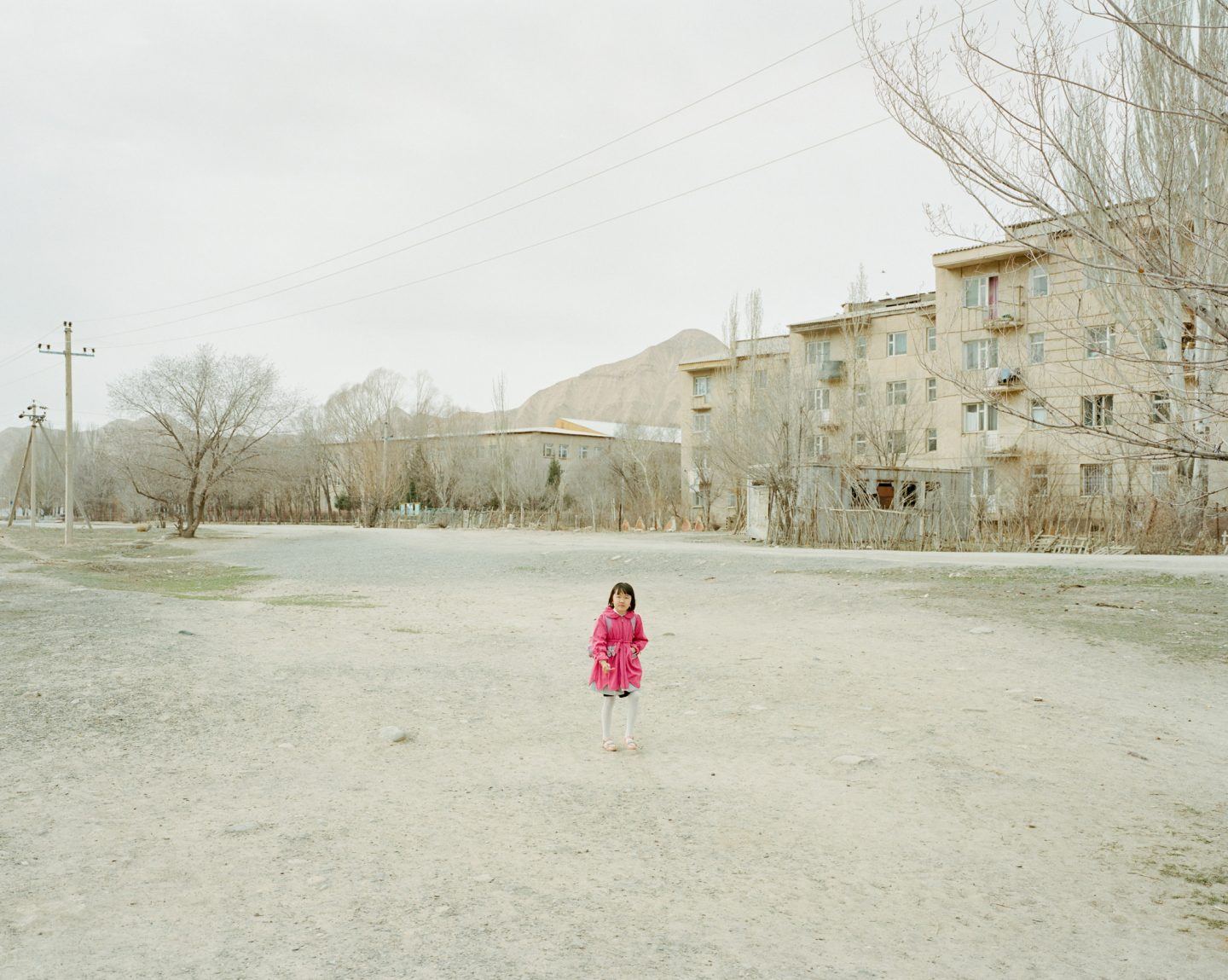Horsehead Nebula by Matthieu Litt — A young child in a bright pink dress stands alone in an expansive dirt courtyard, dwarfed by Soviet-era residential blocks and bare poplar trees.
