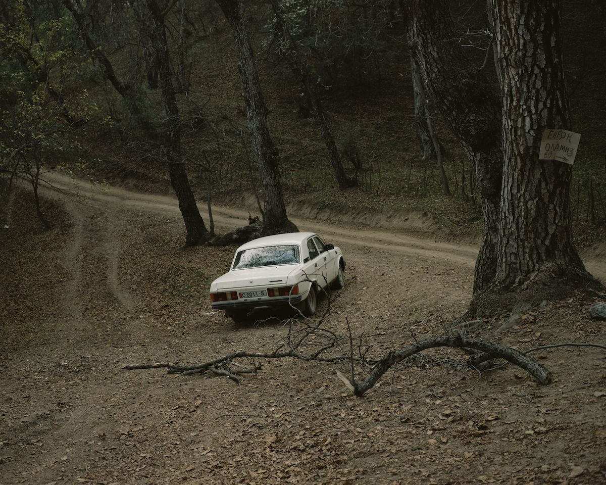 Horsehead Nebula by Matthieu Litt — A white sedan with visible damage sits abandoned on a dirt forest road in Faristan, surrounded by tall trees with grey bark and sparse undergrowth.
