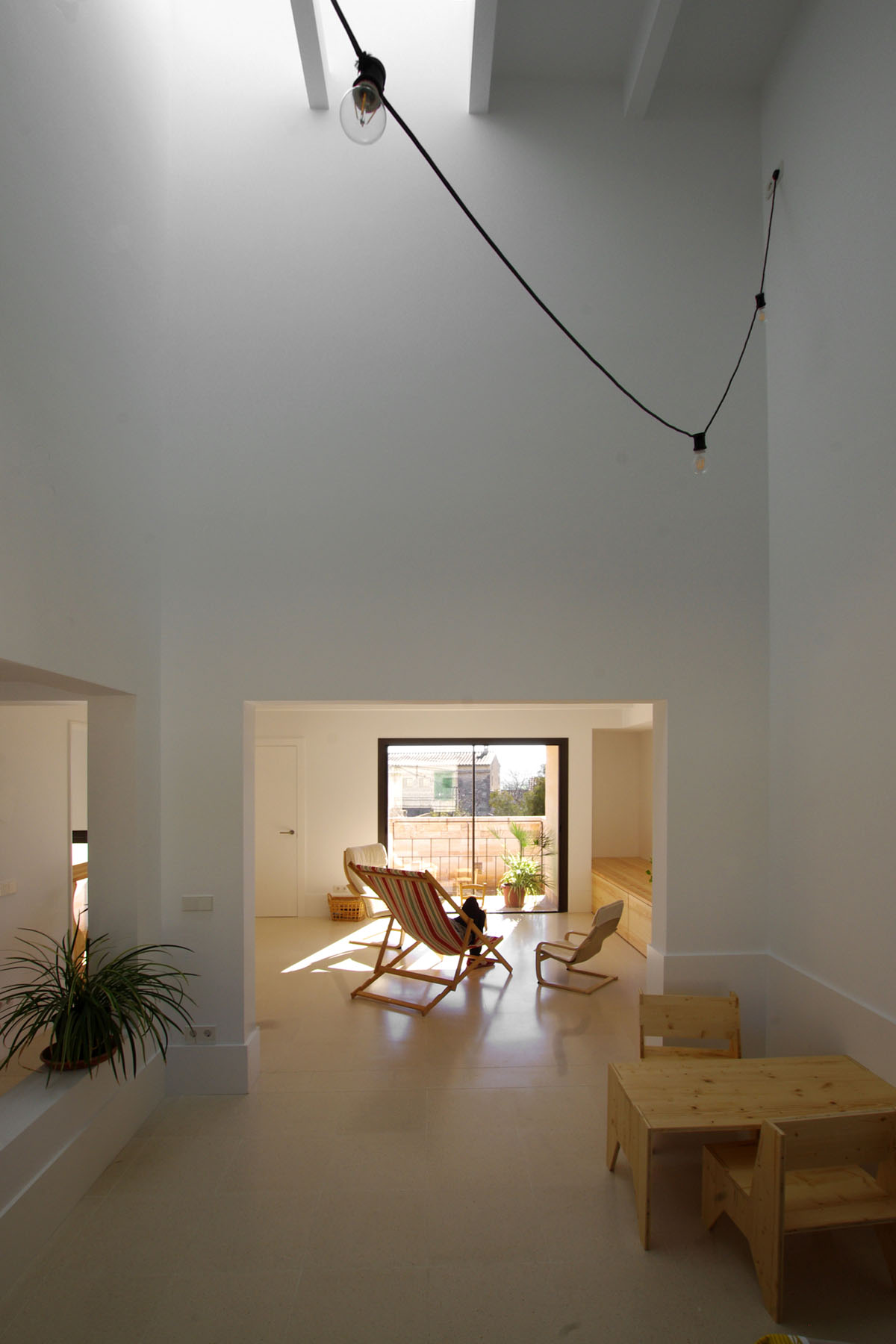 Jordi and África's House by TEd’A arquitectes — A double-height interior space in pale sandstone ashlar, photographed from an upper mezzanine level looking toward glazed doors opening onto a terrace.