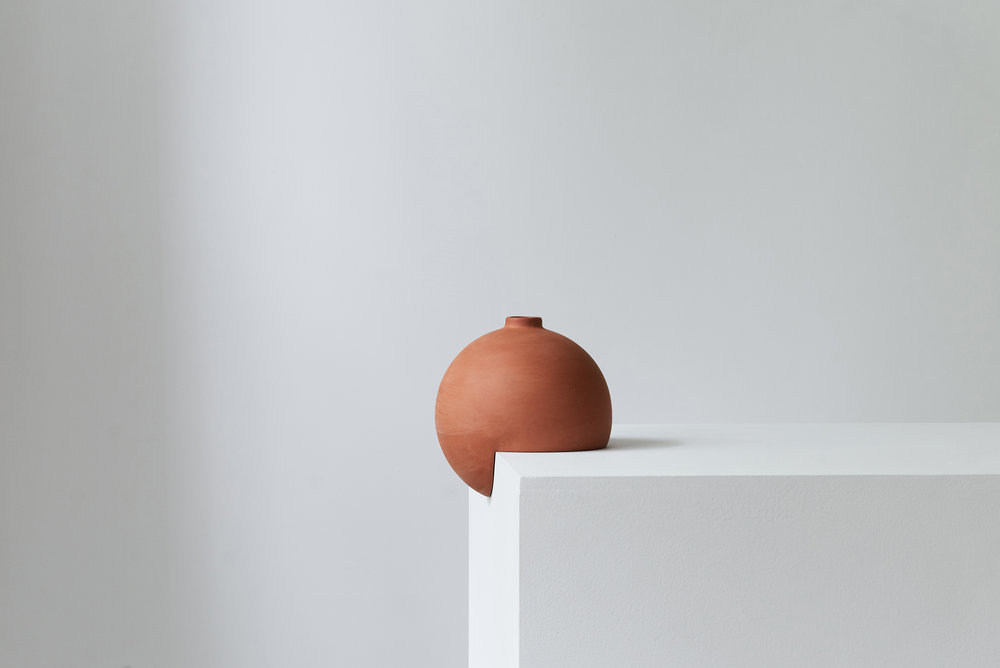 Tumble by Falke Svatun — A terracotta slip-cast stoneware vessel with matte finish sits perched on the edge of a white plaster platform, its spherical form interrupted by a...