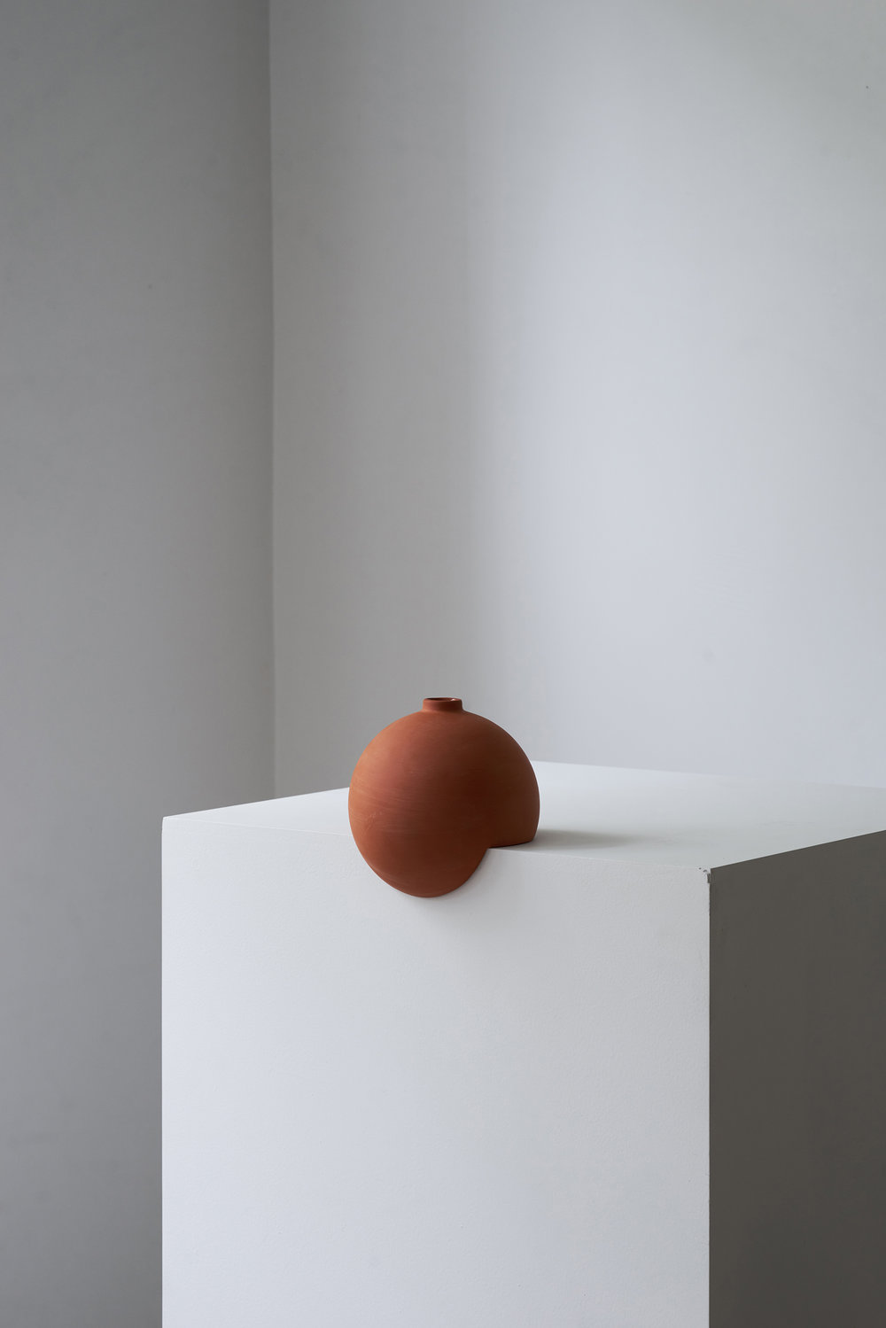 Tumble by Falke Svatun — A terracotta slip-cast stoneware vessel with matte finish sits centered on a white plaster platform, positioned against a gradient gray wall.