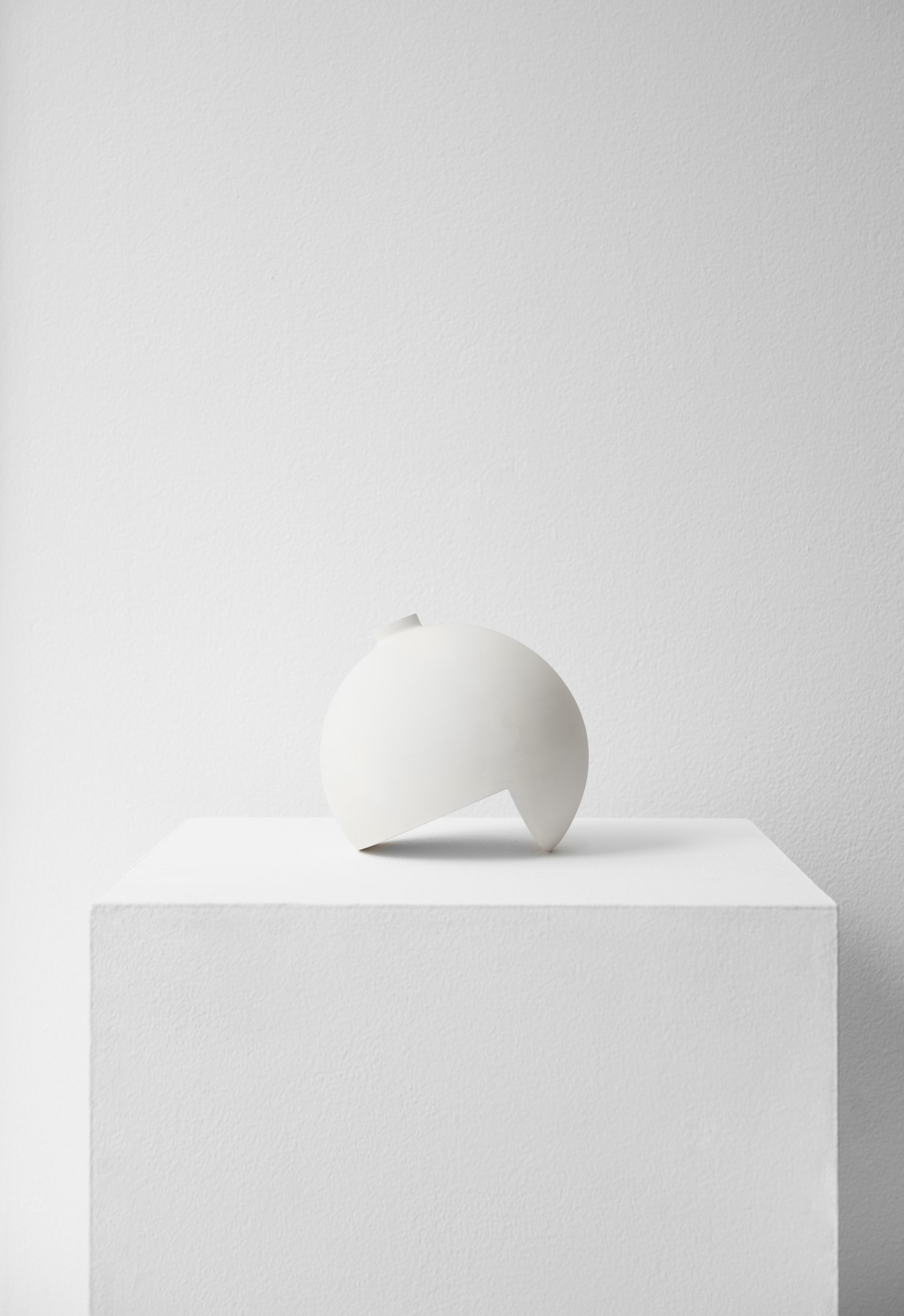 Tumble by Falke Svatun — A cream-colored slip-cast stoneware vessel sits atop a white plaster platform, its spherical form interrupted by a distinctive semicircular cutout at the base.