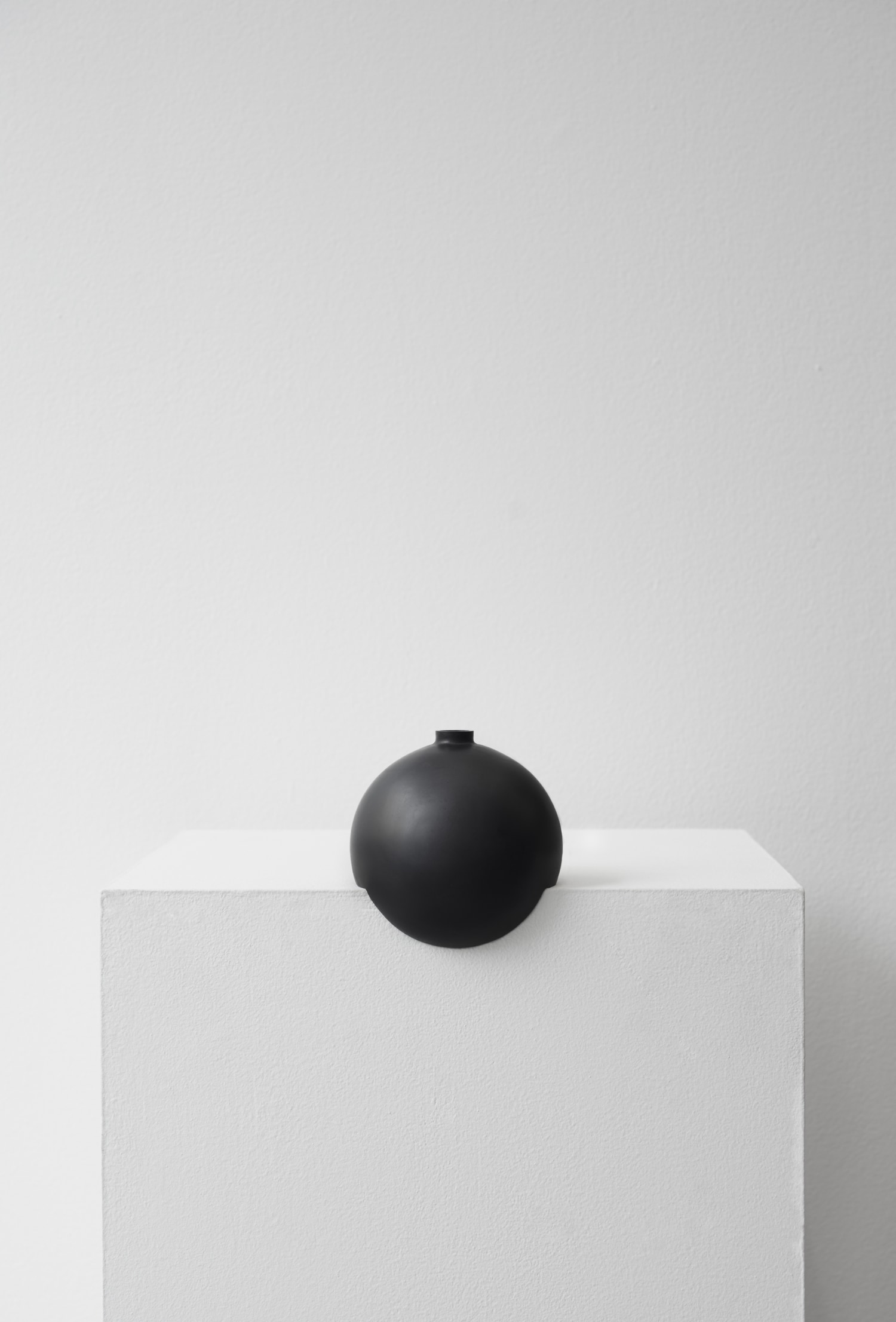Tumble by Falke Svatun — A single black slip-cast stoneware vessel with matte finish sits centered atop a white plaster platform.