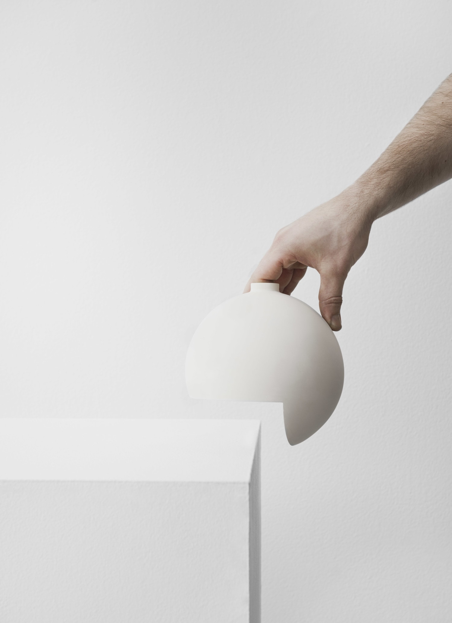 Tumble by Falke Svatun — A hand holds a cream slip-cast stoneware vessel against a white plaster wall, revealing the semicircular cutout at its base—the defining feature enabling...