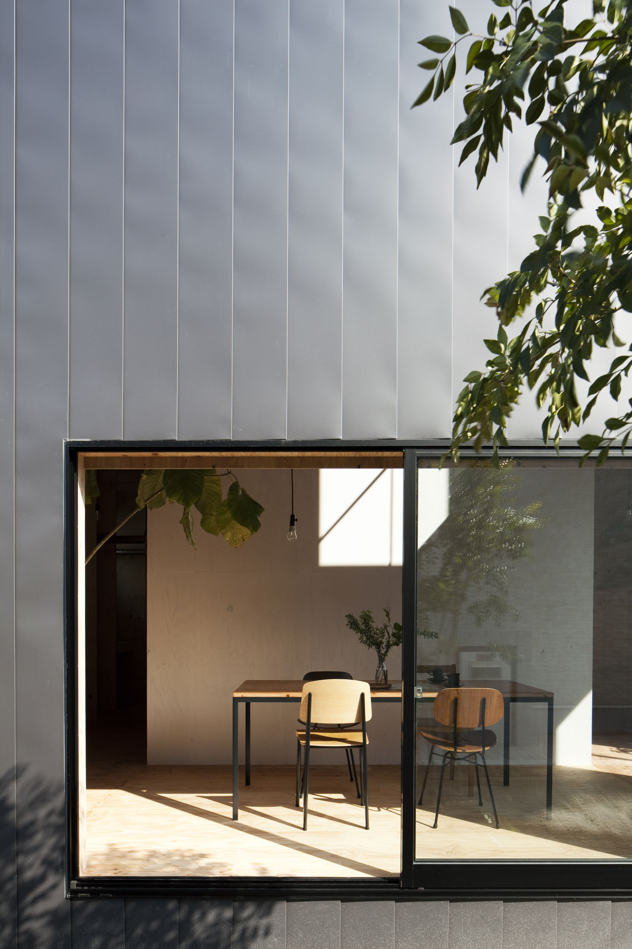 Ant-house by mA-style Architects — A interior view through floor-to-ceiling black-framed glazing reveals a minimalist workspace defined by warm plywood surfaces and natural light.