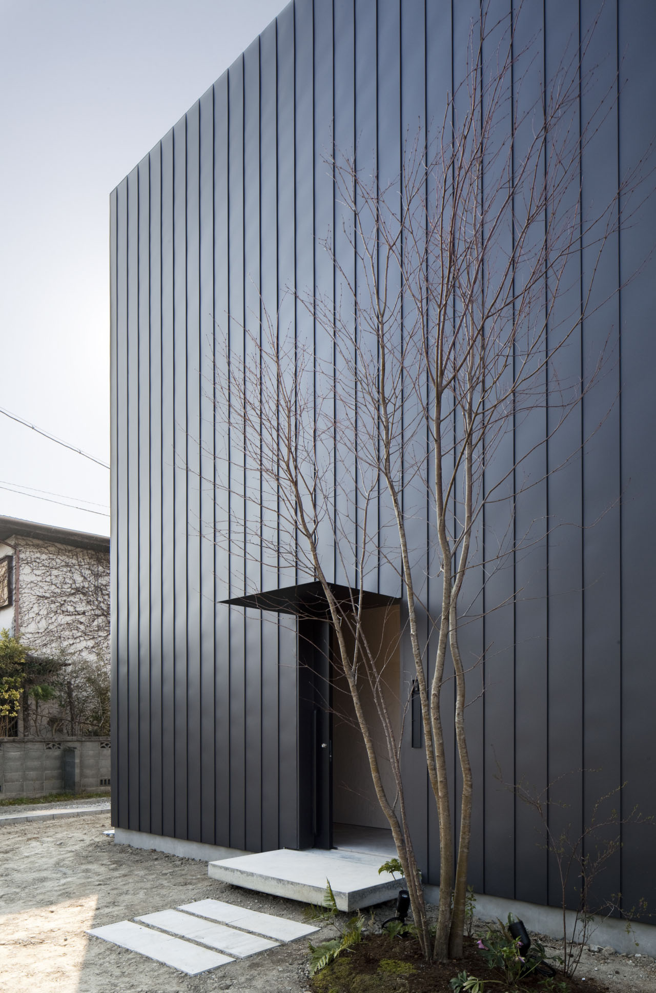 Ant-house by mA-style Architects — The black metal-clad facade of Ant-house presents an unbroken vertical plane interrupted only by a recessed entrance with cantilevered overhang.