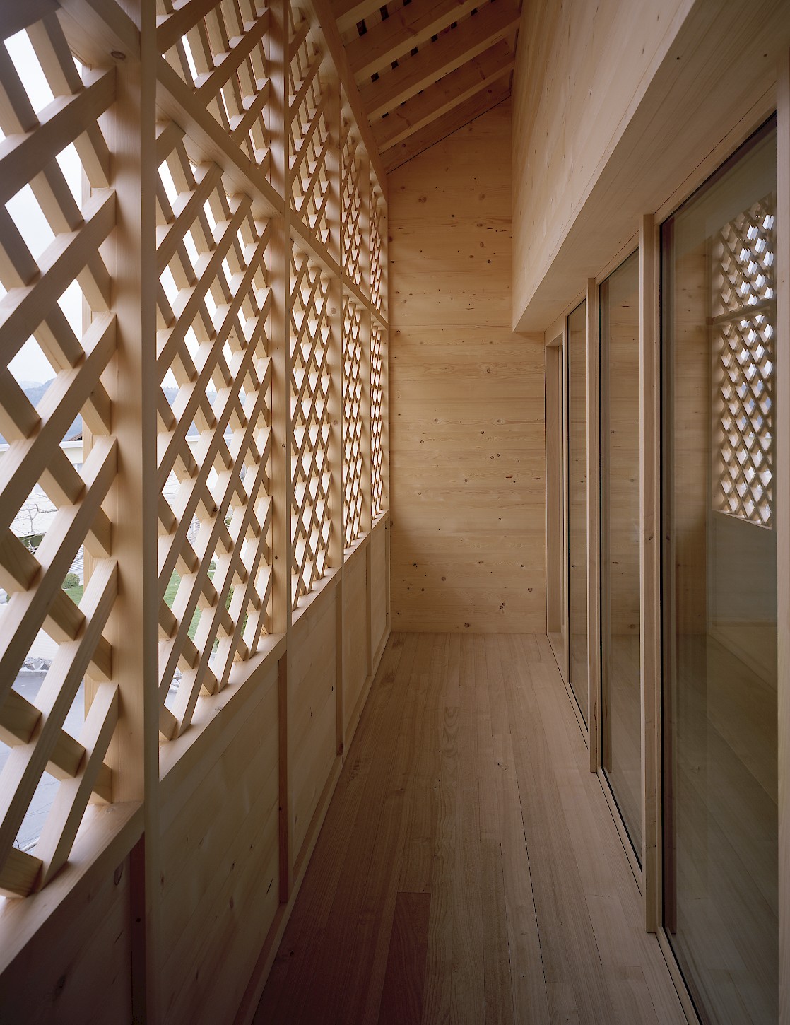 House K Alpnach by Seilerlinhart — A timber-lined corridor extends toward glazed doors, framed by a geometric lattice screen on the left that filters light into a diamond-pattern shadow.