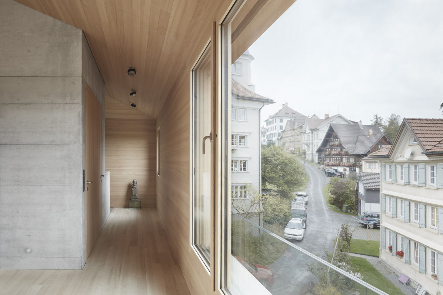 House at the Schopfacker by Bernardo Bader Architects — A timber-lined corridor frames the view toward a Swiss village street through expansive floor-to-ceiling glazing.