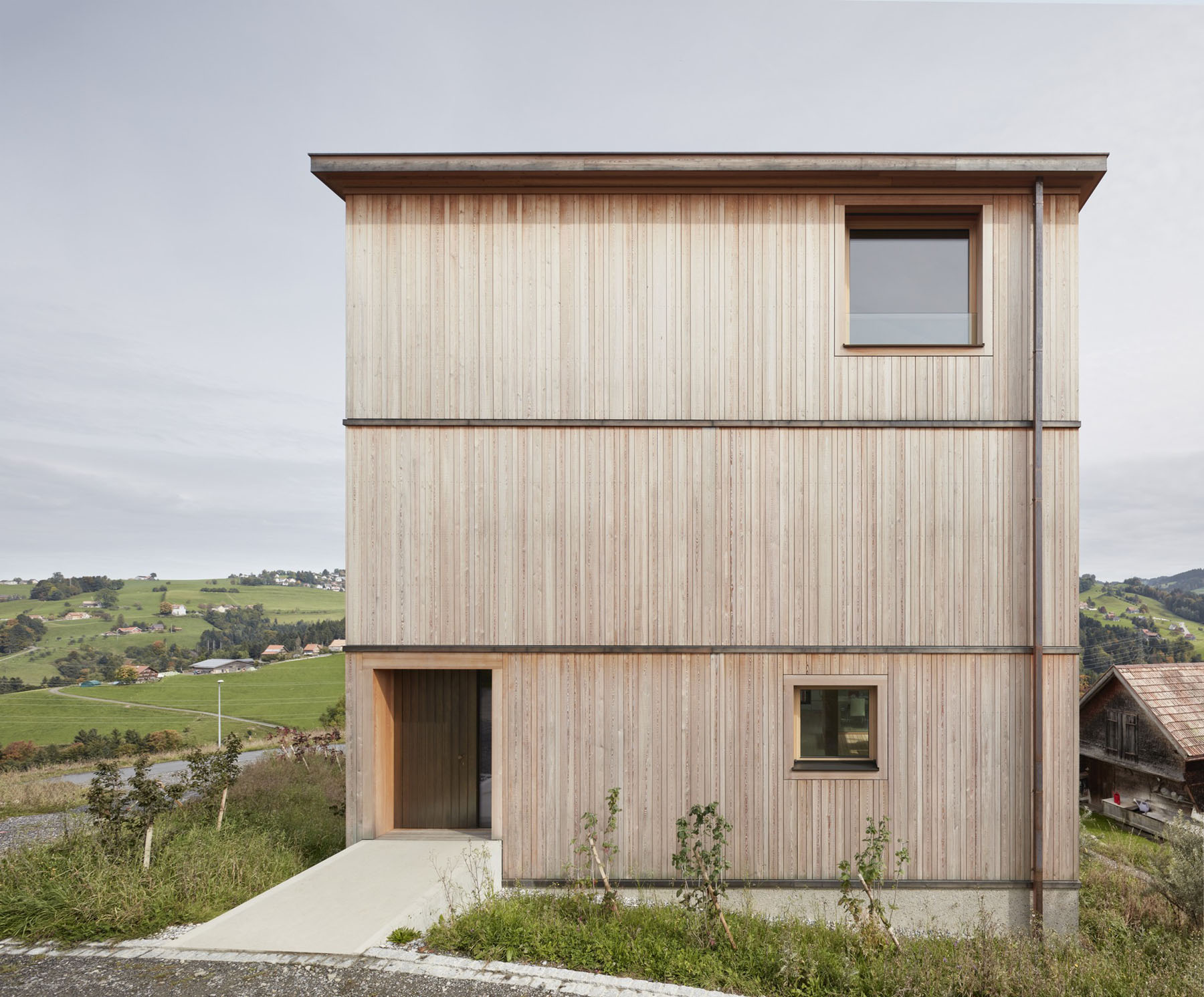 House at the Schopfacker by Bernardo Bader Architects — The timber-clad facade dominates this front elevation photograph, with continuous vertical wood slat cladding punctuated by a single upper window and a...