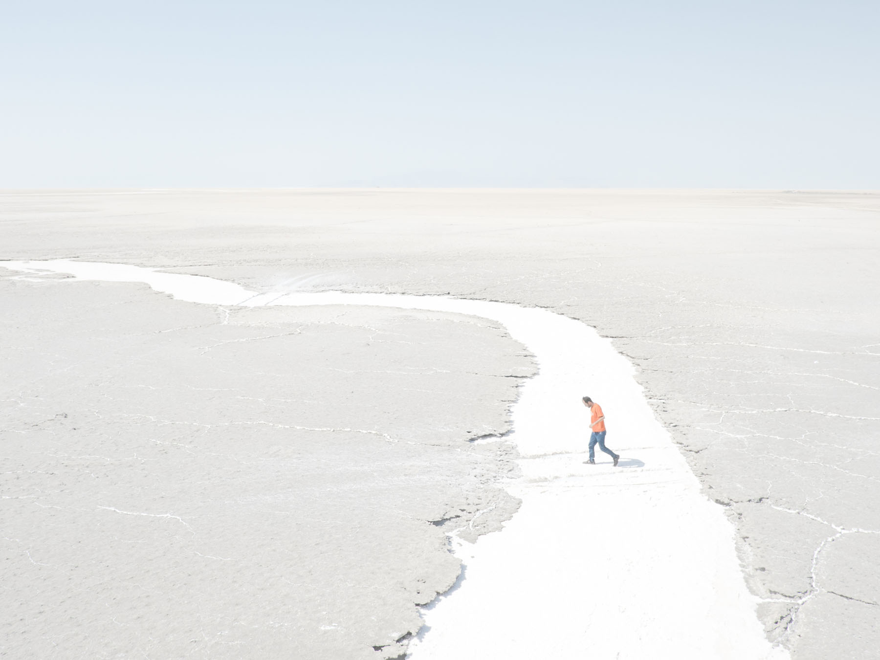 Fading Flamingos by Maximilian Mann — A solitary figure in a bright orange jacket traverses a winding channel of white salt deposits cutting through the grey-beige salt flat of Lake Urmia.