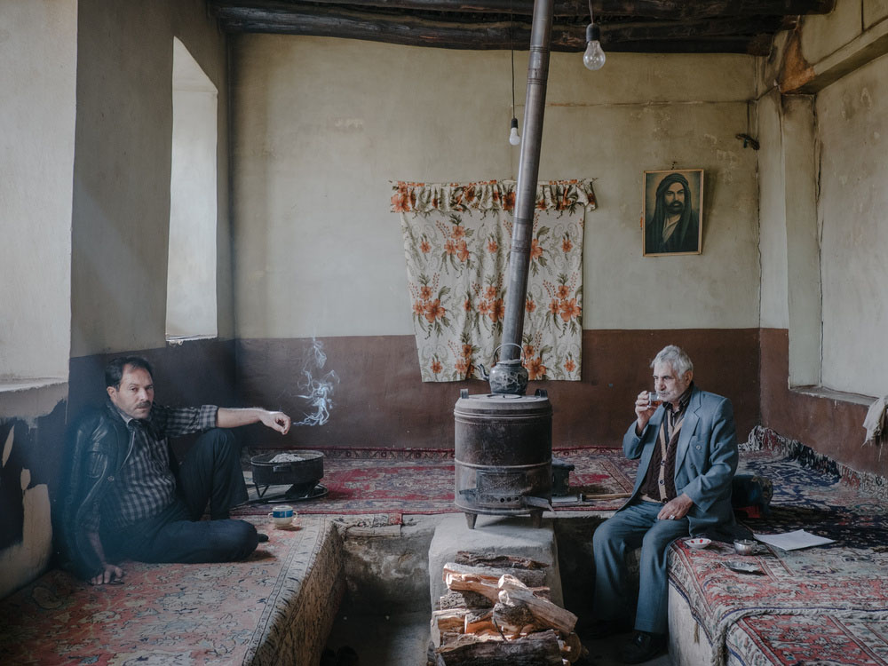 Fading Flamingos by Maximilian Mann — Two men sit cross-legged on worn Persian rugs in a sparse interior, separated by a central stove with metal piping.