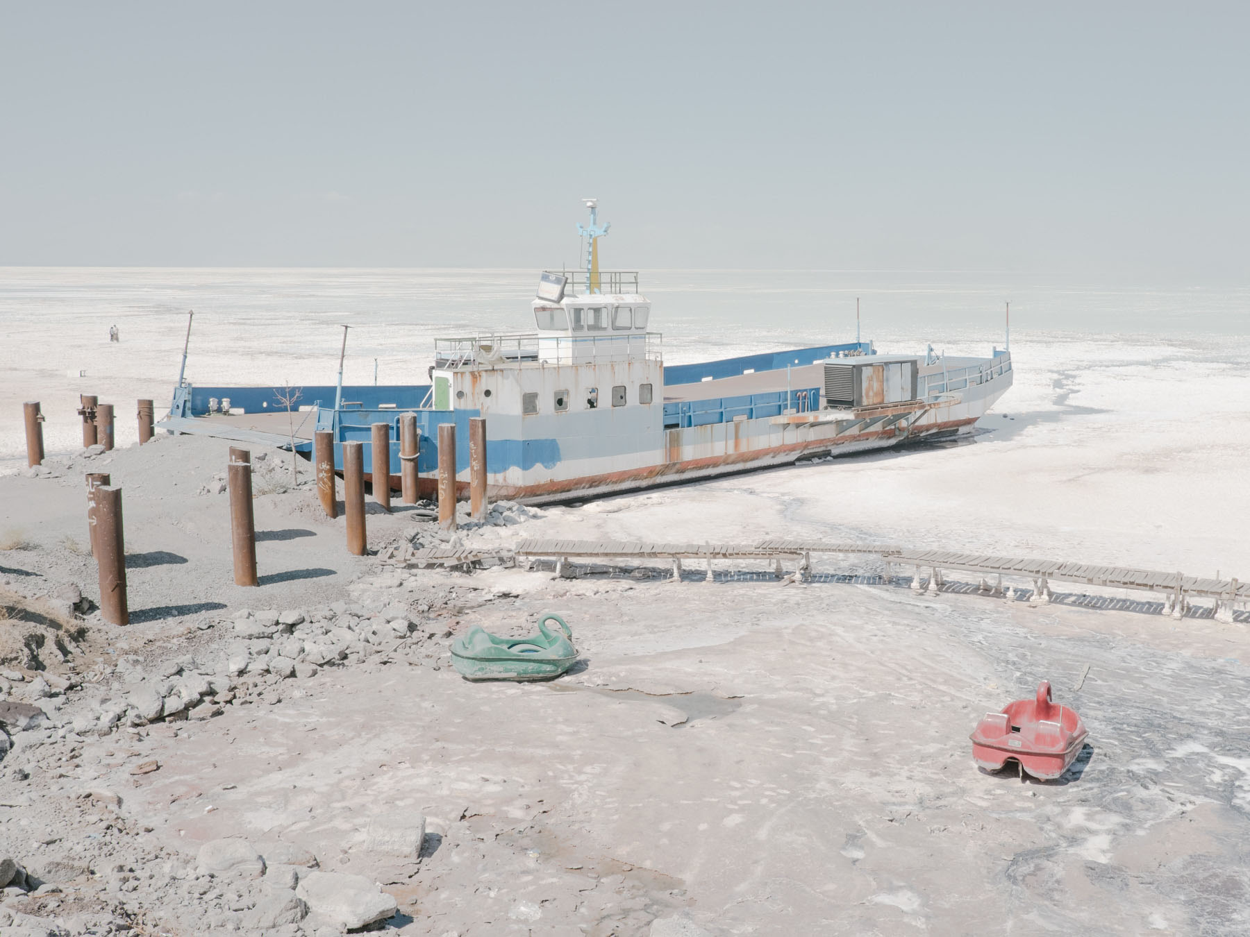 Fading Flamingos by Maximilian Mann — A stranded commercial fishing vessel rests on the desiccated bed of Lake Urmia, its blue and white hull listing against wooden pilings driven into...