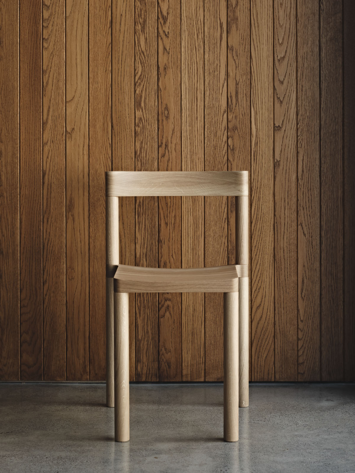 Pier Chair by Leonard Kadid — The Pier Chair by Leonard Kadid is photographed front-on against a darkened oak plank backdrop, its light ash-stained frame in sharp contrast.