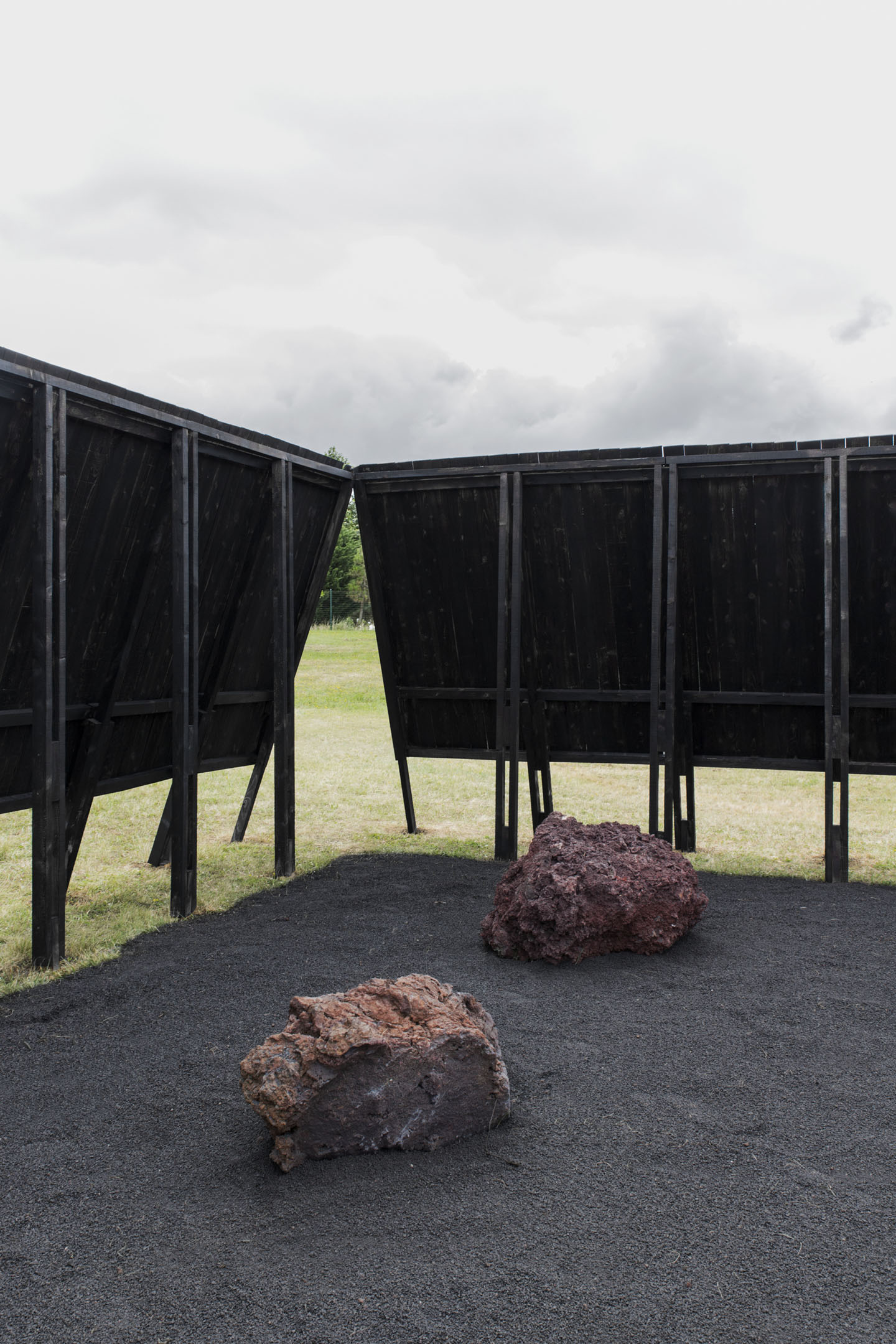 Inbetween Pavilion by Pontoatelier — Two volcanic rocks rest on dark volcanic pavement within the pavilion's interior, their reddish-brown surfaces contrasting against the charred timber structure.
