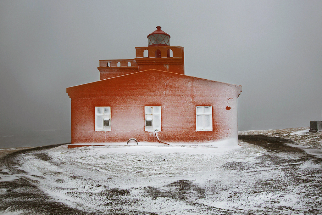 Snjór by Christophe Jacrot — A rust-colored brick lighthouse structure stands isolated on snow-covered volcanic terrain in Iceland.