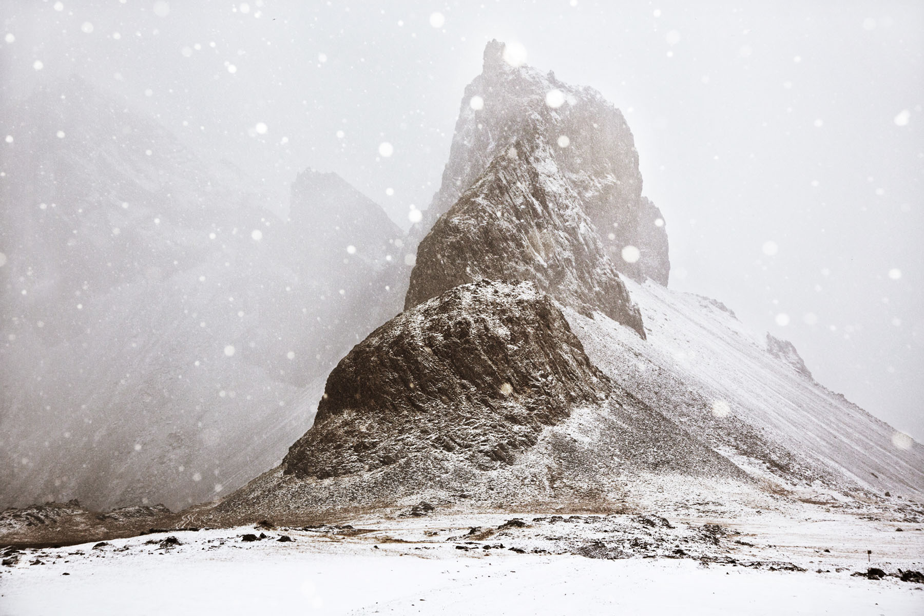 Snjór by Christophe Jacrot — A monolithic rock formation rises steeply from a snow-covered plain in heavy snowfall, its dark volcanic stone face creating stark contrast against the...