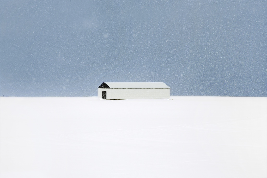 Snjór by Christophe Jacrot — A solitary structure sits centered on a snow-covered plateau in Iceland, photographed during active snowfall.