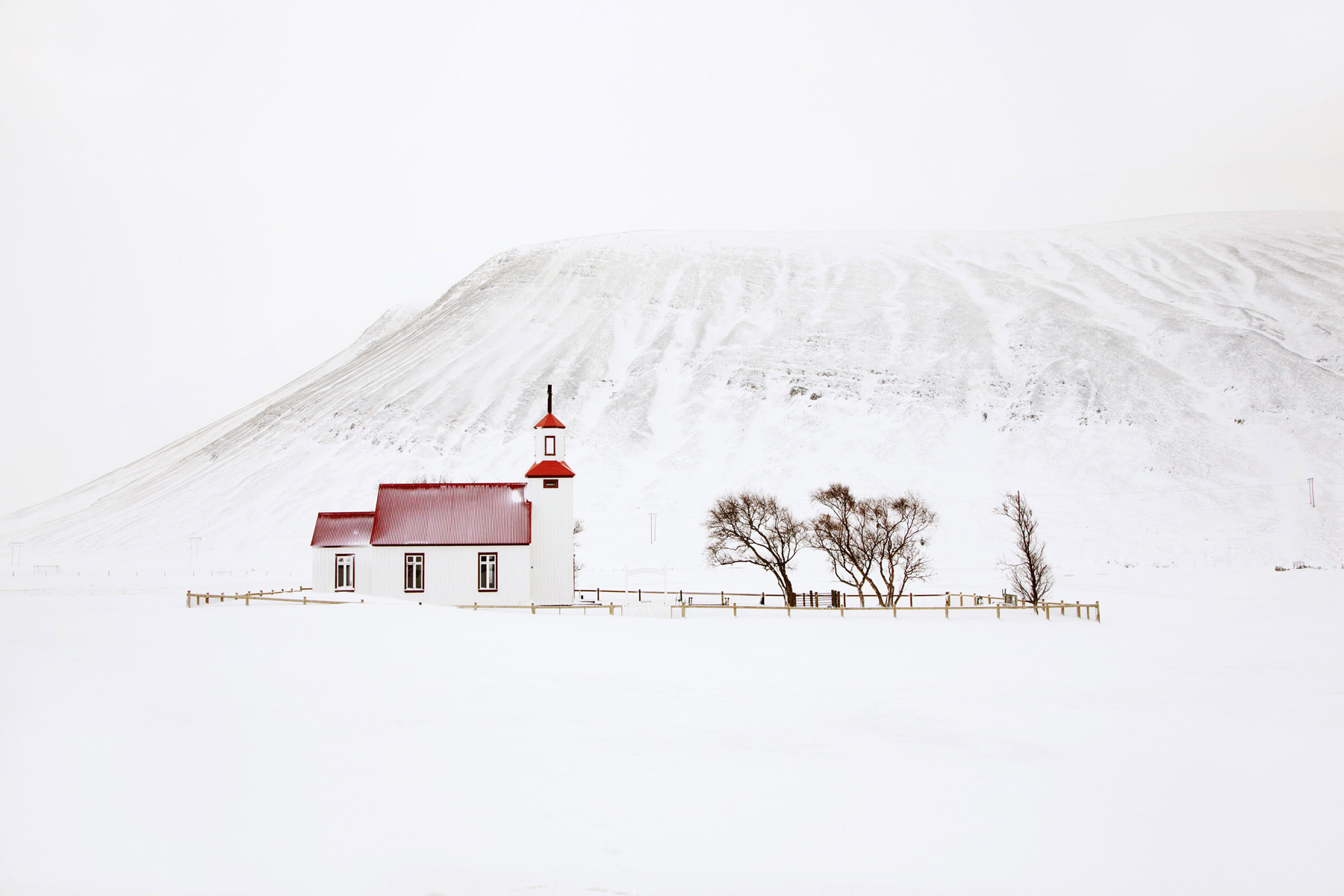 Snjór by Christophe Jacrot — A wooden church with red corrugated-metal roof and white walls stands isolated against a snow-covered volcanic mountainside in Iceland.