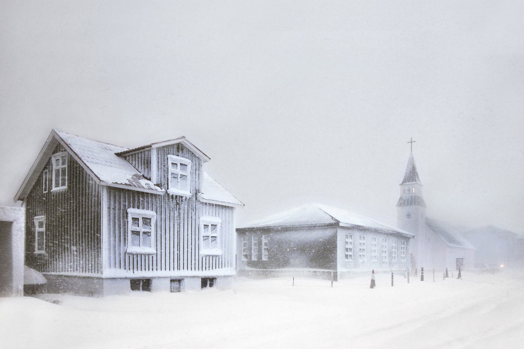 Snjór by Christophe Jacrot — A corrugated-metal church dominates the foreground of this snow-laden Icelandic settlement, its pitched roof and white-trimmed windows contrasting sharply...