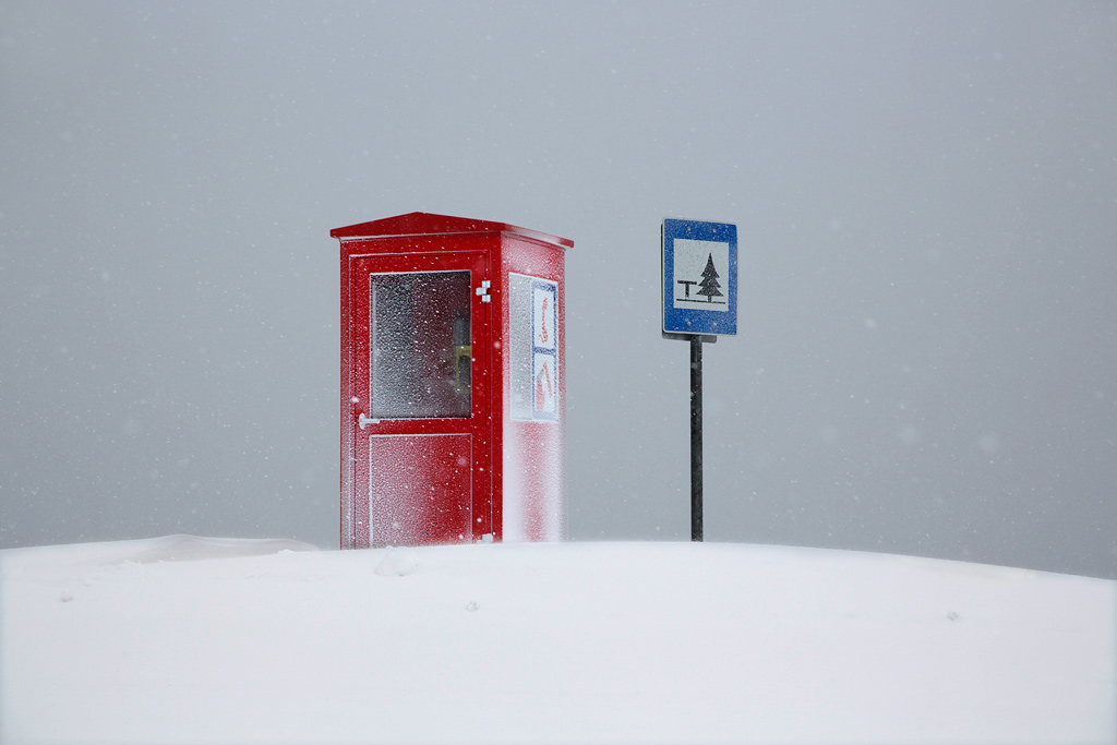 Snjór by Christophe Jacrot — A red Swiss telephone booth stands isolated in a snow-covered landscape, its geometric form reinforced by a white cross emblem on the door.