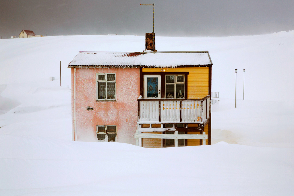 Snjór by Christophe Jacrot — A small residential structure emerges from deep snow in Iceland's interior, its colorful facades—pink brick, yellow timber cladding, and ochre trim—creating...