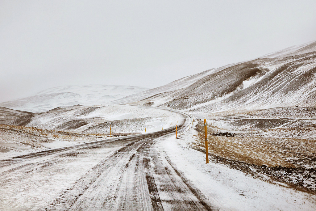 Snjór by Christophe Jacrot — A snow-covered road in Iceland curves through volcanic terrain, marked by yellow reflective posts at regular intervals.