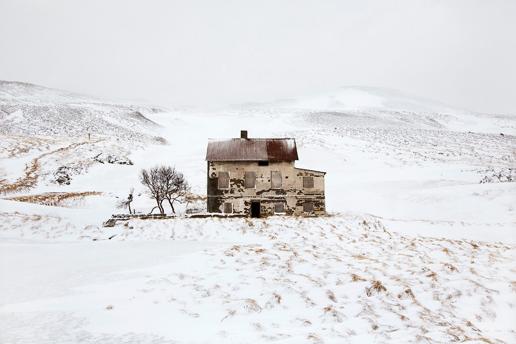 Snjór by Christophe Jacrot — A deteriorating two-storey timber-frame structure with corrugated metal roof stands isolated in a snow-covered volcanic landscape.