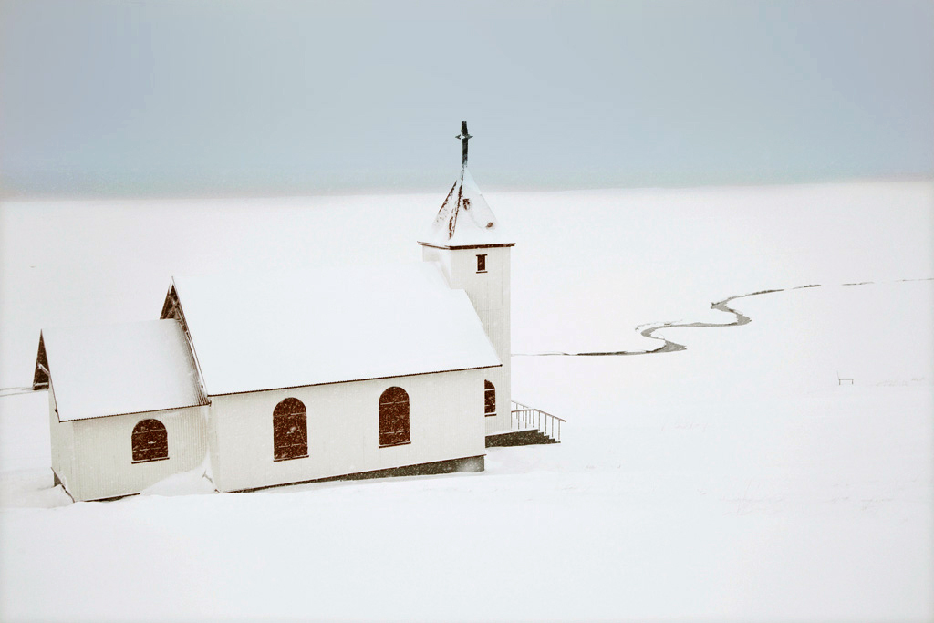 Snjór by Christophe Jacrot — A white wooden chapel with pitched roof and cross-topped steeple stands isolated across snow-covered terrain in Iceland.