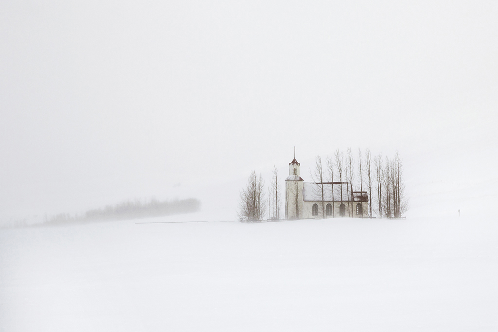 Snjór by Christophe Jacrot — A solitary timber-frame church with a pitched roof and steeple emerges through heavy snowfall on an Icelandic plateau.