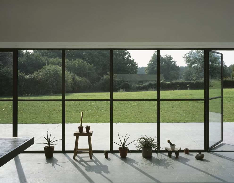 Stable Acre House by David Kohn — Interior threshold framed by dark-profile steel glazing, with potted plants and a wooden easel casting sharp shadows across light concrete flooring.