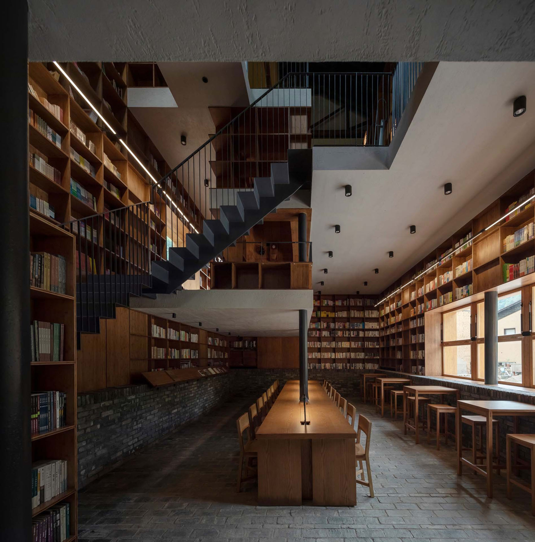 Capsule Hotel and Bookstore in Village Qinglongwu by Atelier tao+c — A linear interior perspective reveals a two-story library and bookstore with timber shelving systems running the full height of both walls.