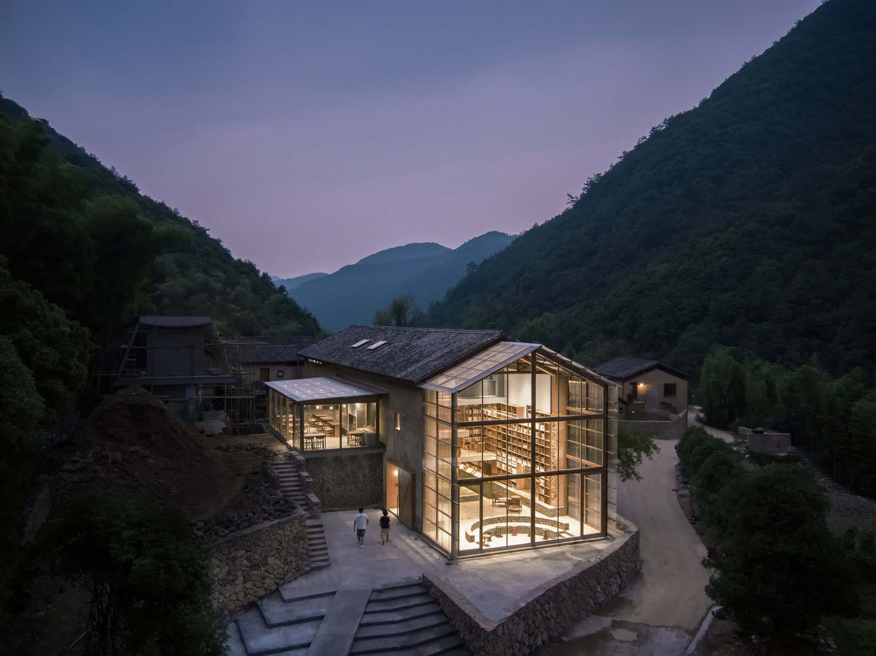Capsule Hotel and Bookstore in Village Qinglongwu by Atelier tao+c — A newly inserted modernist volume with floor-to-ceiling gridded glazing punctures the historic stone masonry envelope of a village structure nestled in a...