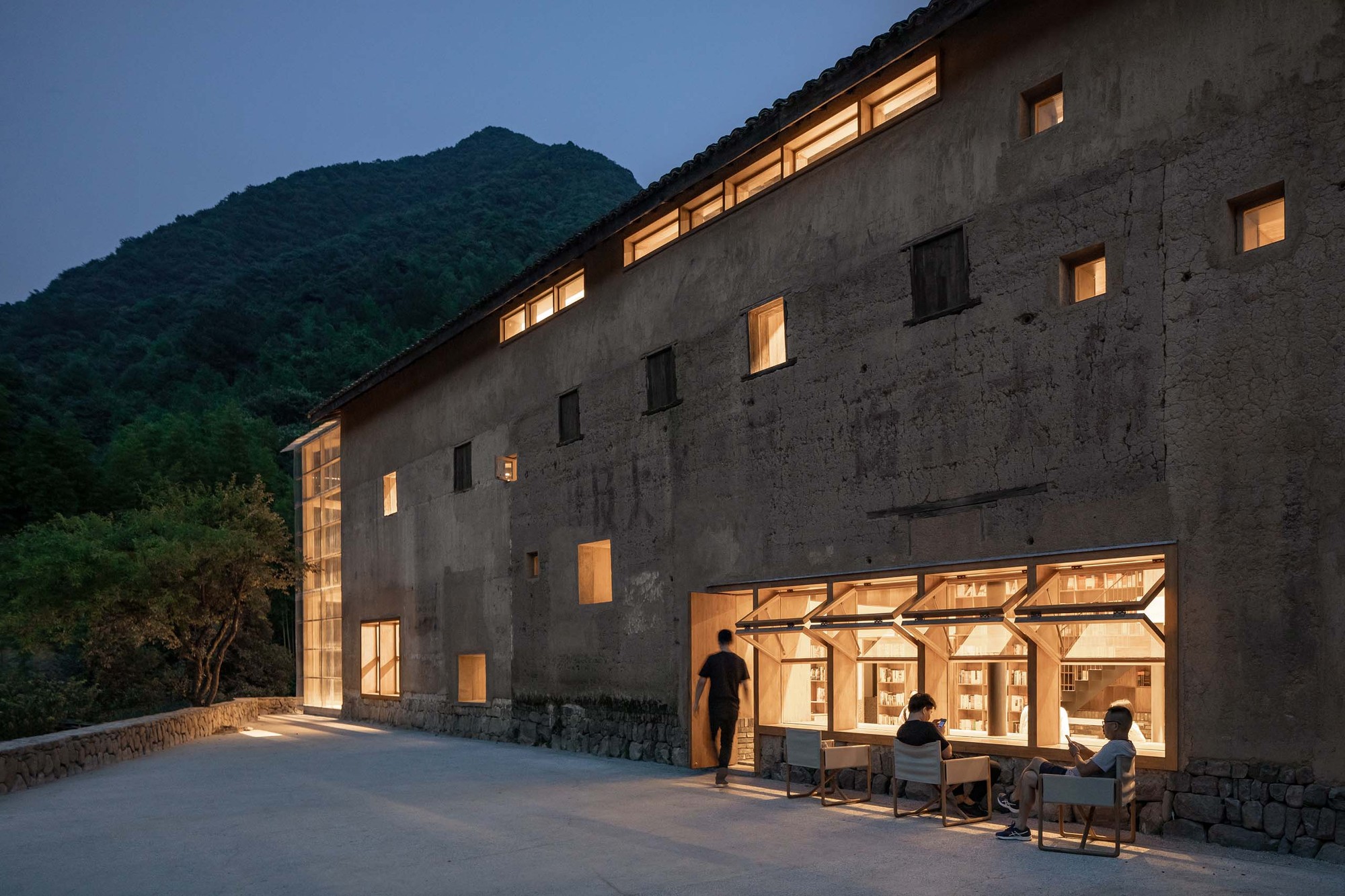 Capsule Hotel and Bookstore in Village Qinglongwu by Atelier tao+c — A renovated village structure at dusk demonstrates adaptive reuse of weathered stone masonry and timber framing.