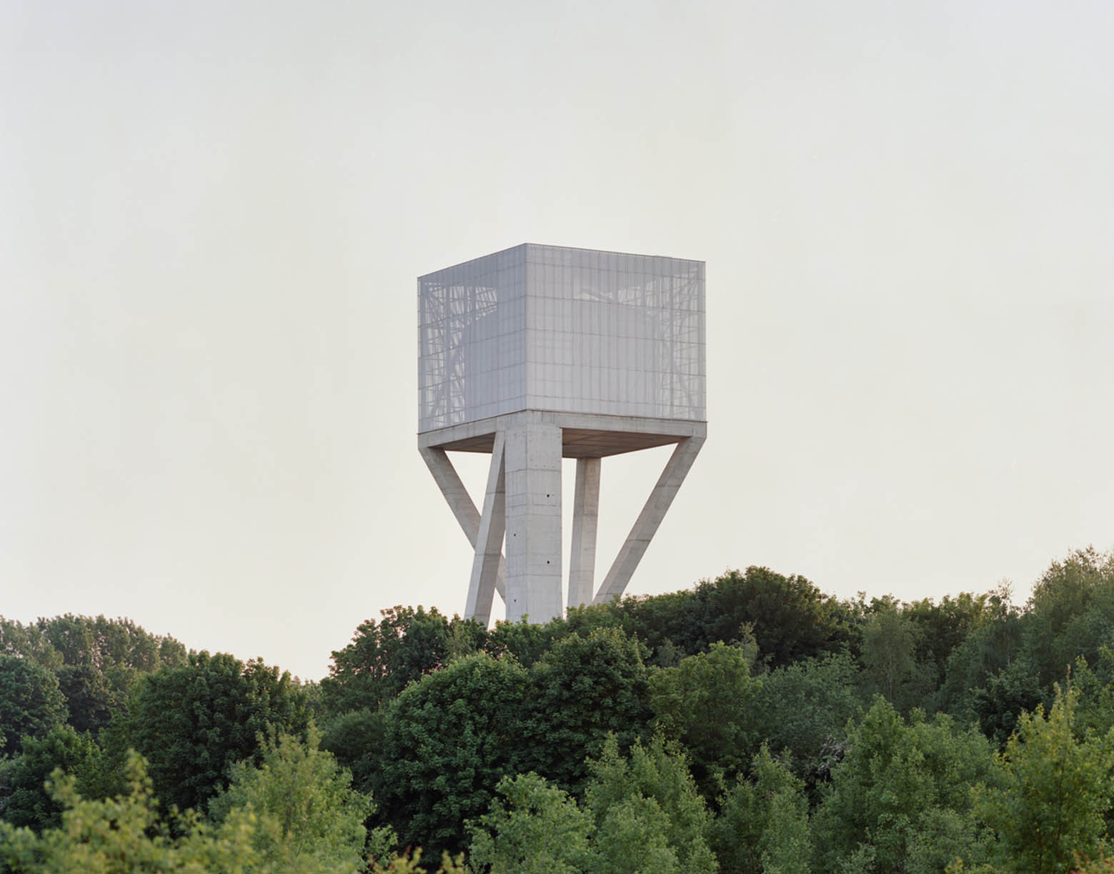 Water Tower by V+ — A cubic concrete water tank elevated on four splayed legs rises above dense forest canopy in Mons, Belgium.