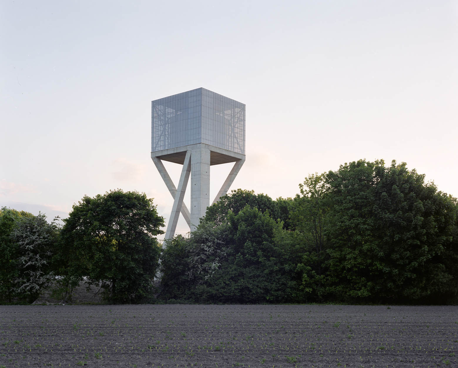 Water Tower by V+ — A cubic water tank elevated on four splayed concrete legs rises above dense woodland, its mesh-clad walls creating visual permeability against the overcast sky.