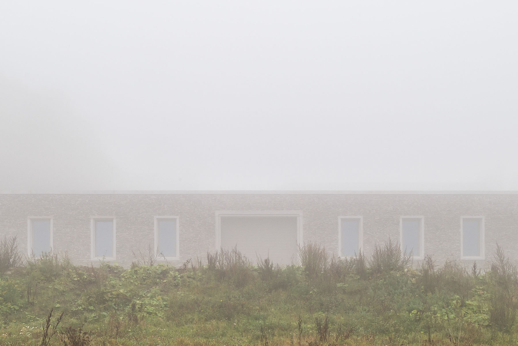Nový Dvůr Industry by John Pawson — The rendered facade of Nový Dvůr emerges from dense foreground vegetation in heavy fog, with six flush rectangular window openings arranged in regular...