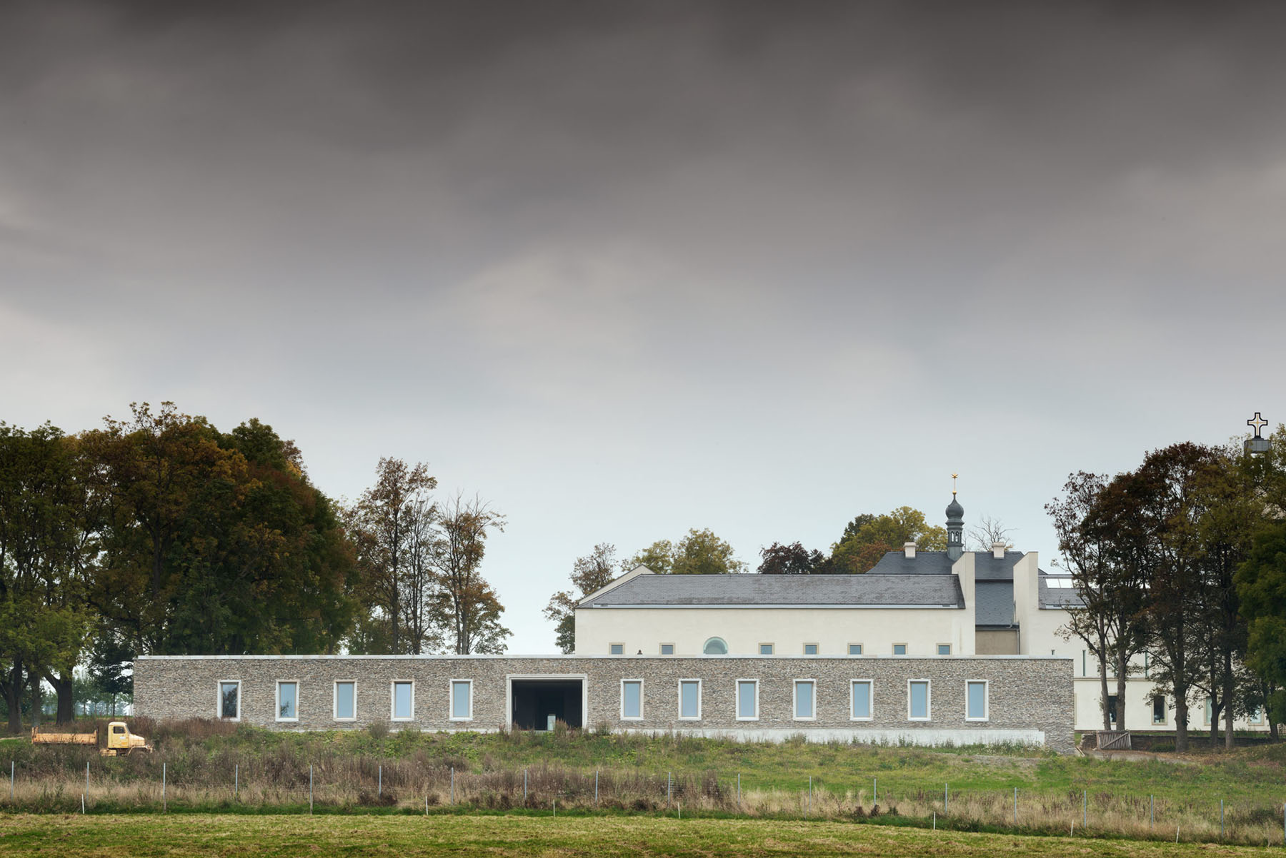 Nový Dvůr Industry by John Pawson — The rendered facade of the industrial workshops appears rendered in stone or aggregate finish, its horizontal bands of flush rectangular windows creating a...