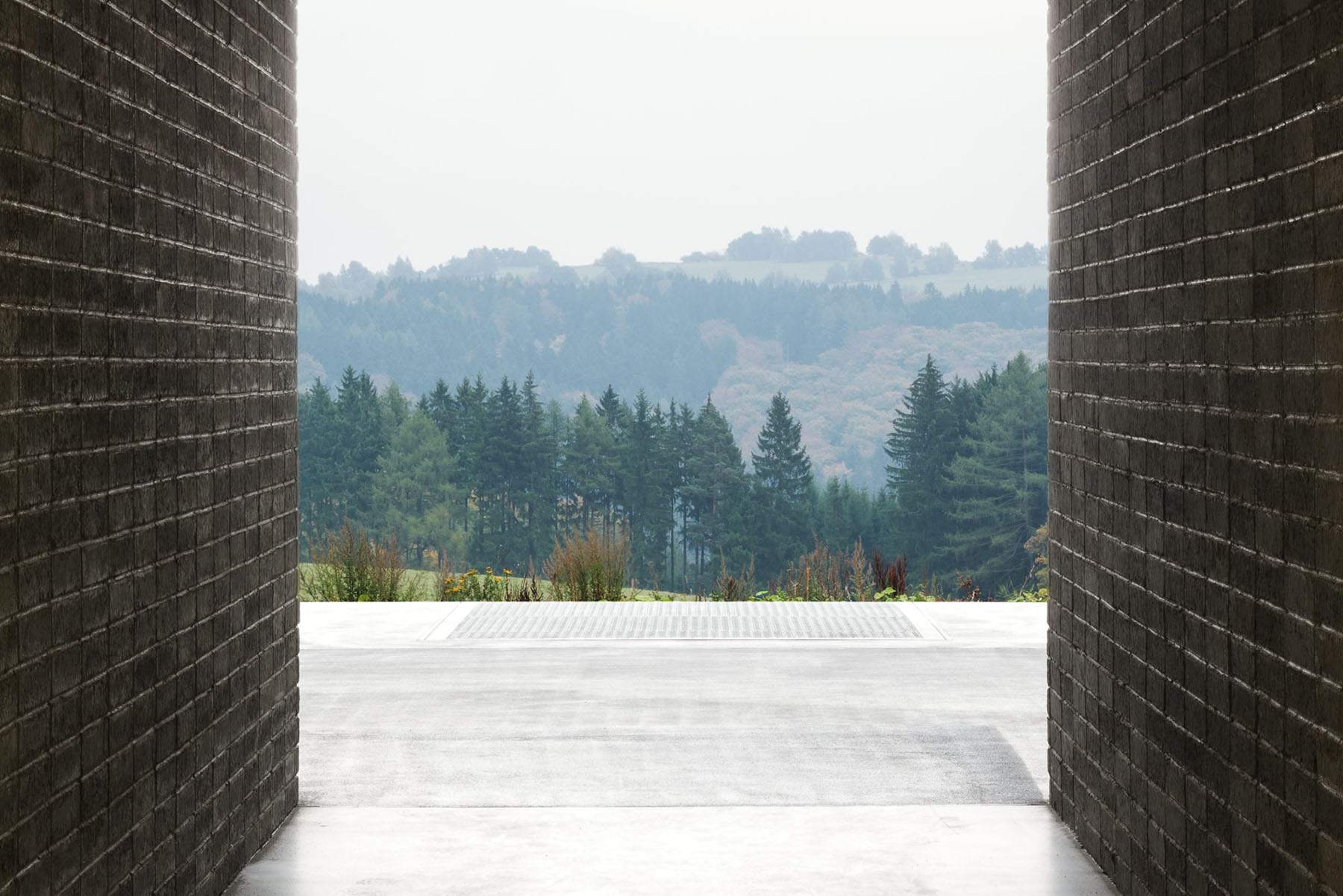 Nový Dvůr Industry by John Pawson — Framed through a passage between two dark brick walls, the industrial workshop reveals a horizontal concrete platform extending toward a forested landscape.