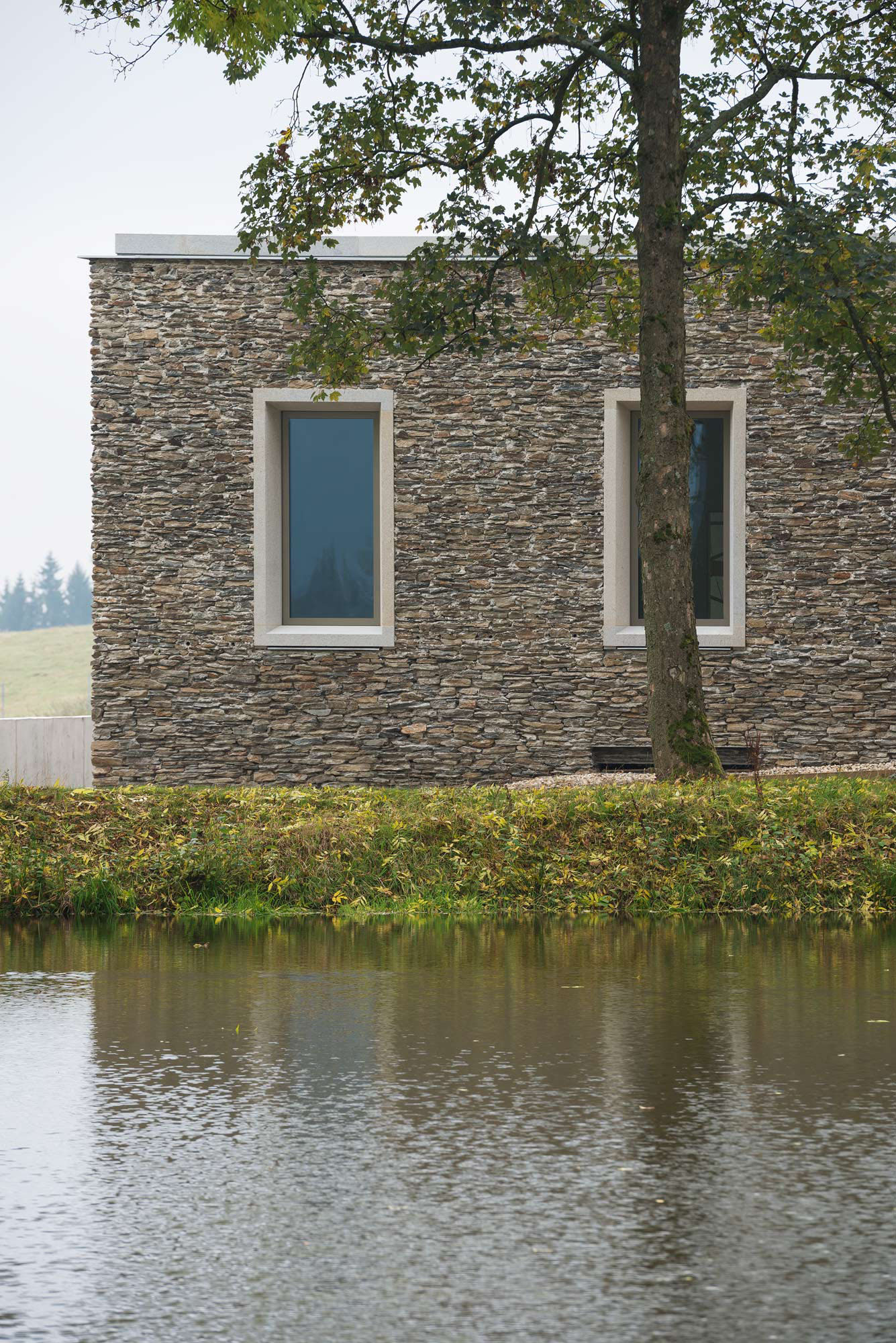Nový Dvůr Industry by John Pawson — A rendered stone facade fronts the water, its horizontal datum marked by two rectangular window openings with cream surrounds set in regular rhythm across...
