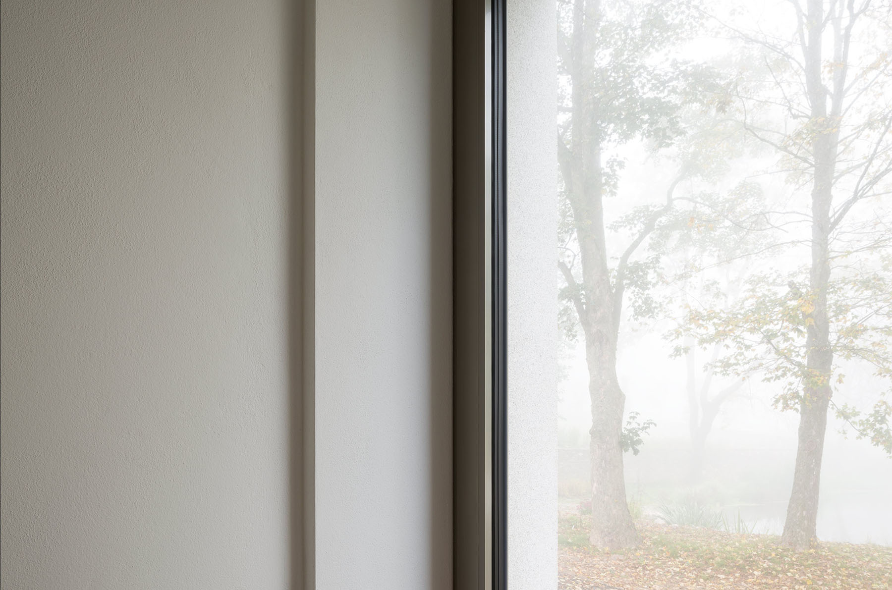 Nový Dvůr Industry by John Pawson — A rendered facade glimpsed through floor-to-ceiling glazing, its cream-colored surface punctuated by rectangular window openings in regular rhythm.