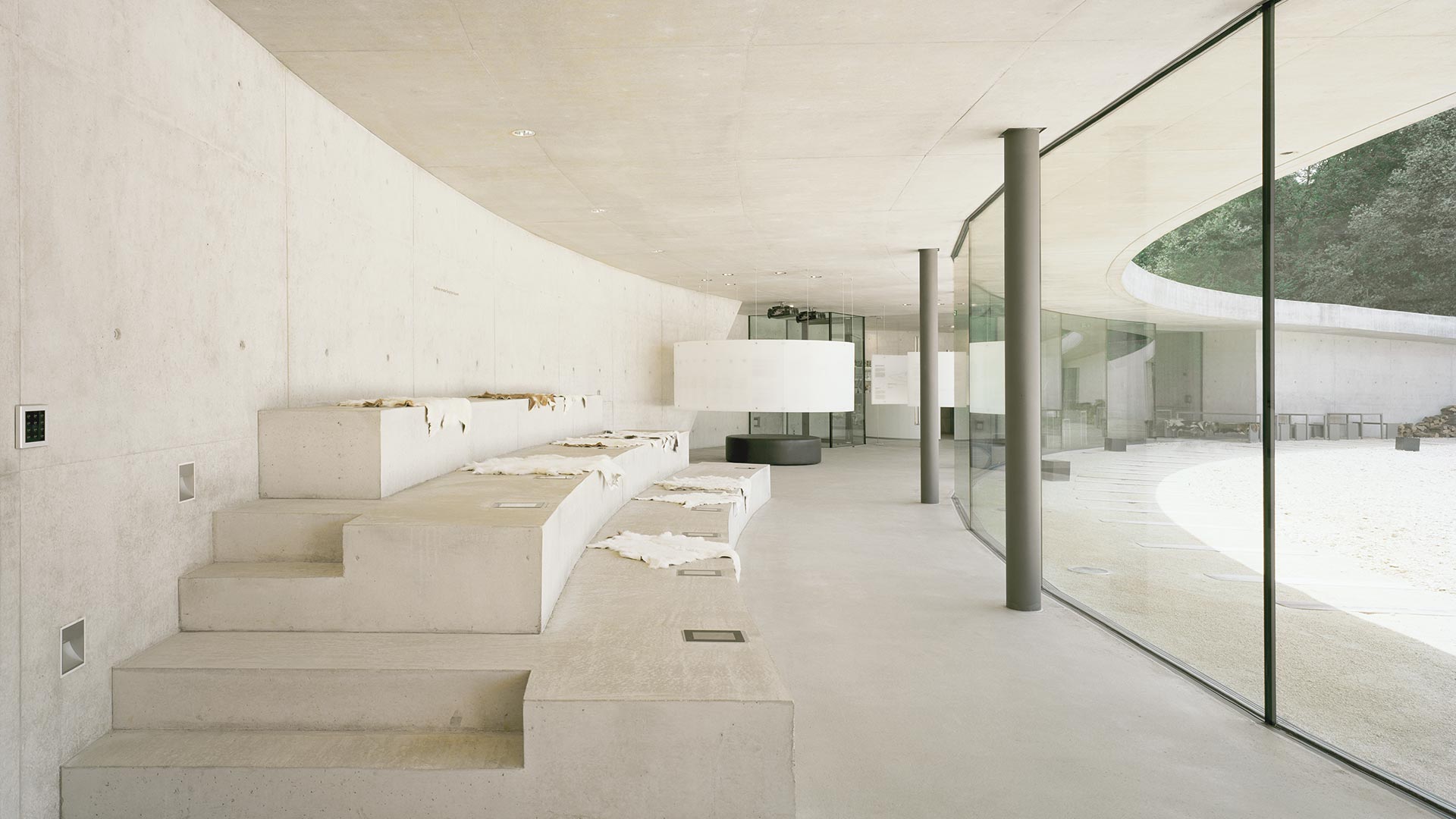 Information Center Archäopark Vogelherd by Ritter Jockisch Architects — Interior view of the glazed foyer revealing tiered concrete platforms ascending leftward, each topped with pale fabric-draped elements.