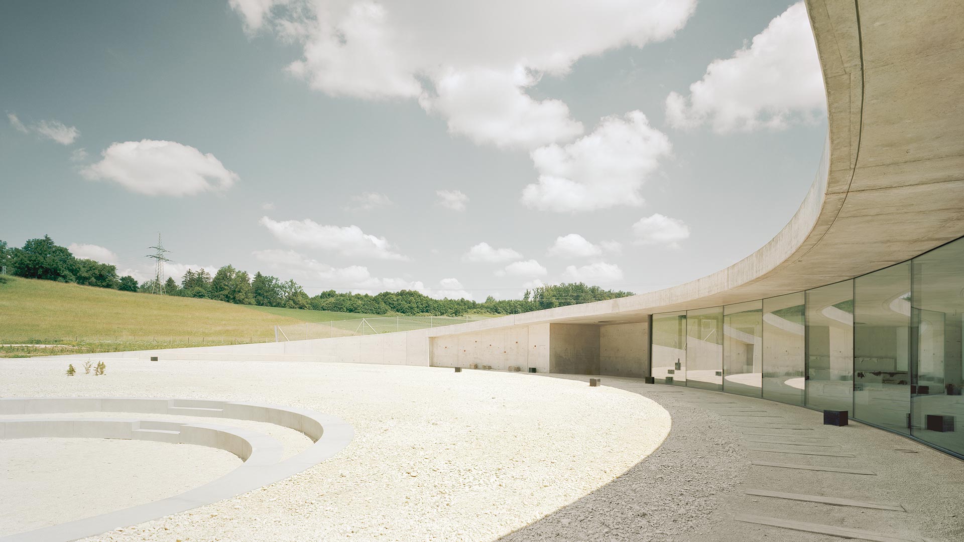 Information Center Archäopark Vogelherd by Ritter Jockisch Architects — The glazed courtyard elevation presents an unobstructed view across gravel and timber decking toward the Vogelherd hill.