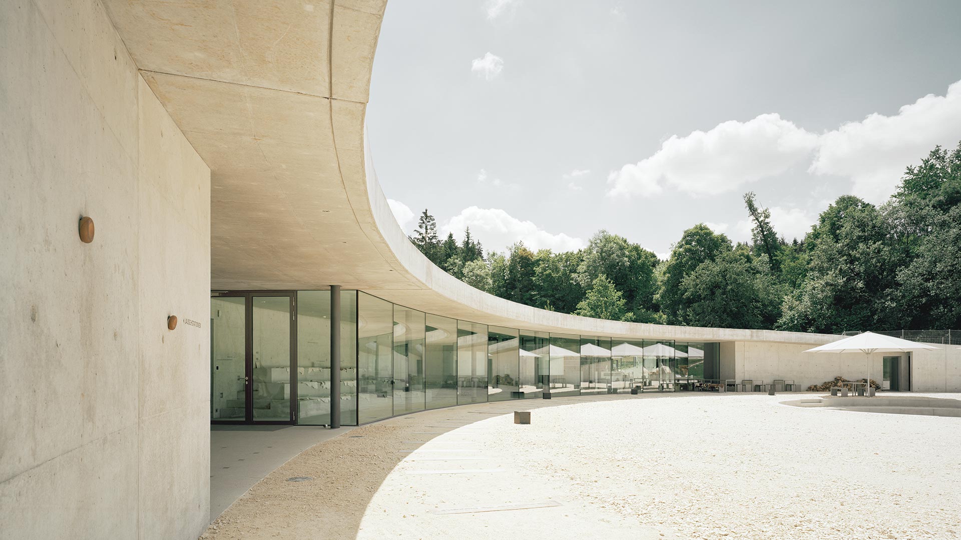Information Center Archäopark Vogelherd by Ritter Jockisch Architects — The courtyard elevation presents a continuous glazed facade spanning the building's full width, framing views of the forested Vogelherd hill beyond.