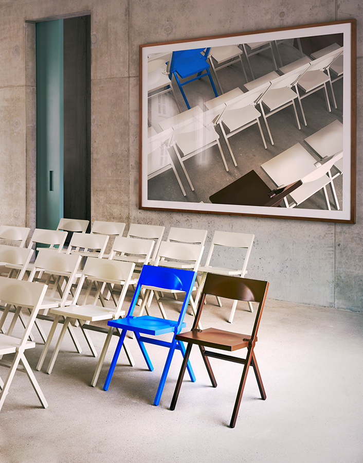 Piana Chair by David Chipperfield — Three Piana chairs occupy a concrete interior: a bright cobalt-blue example positioned prominently in the foreground, accompanied by a bronze-toned variant...