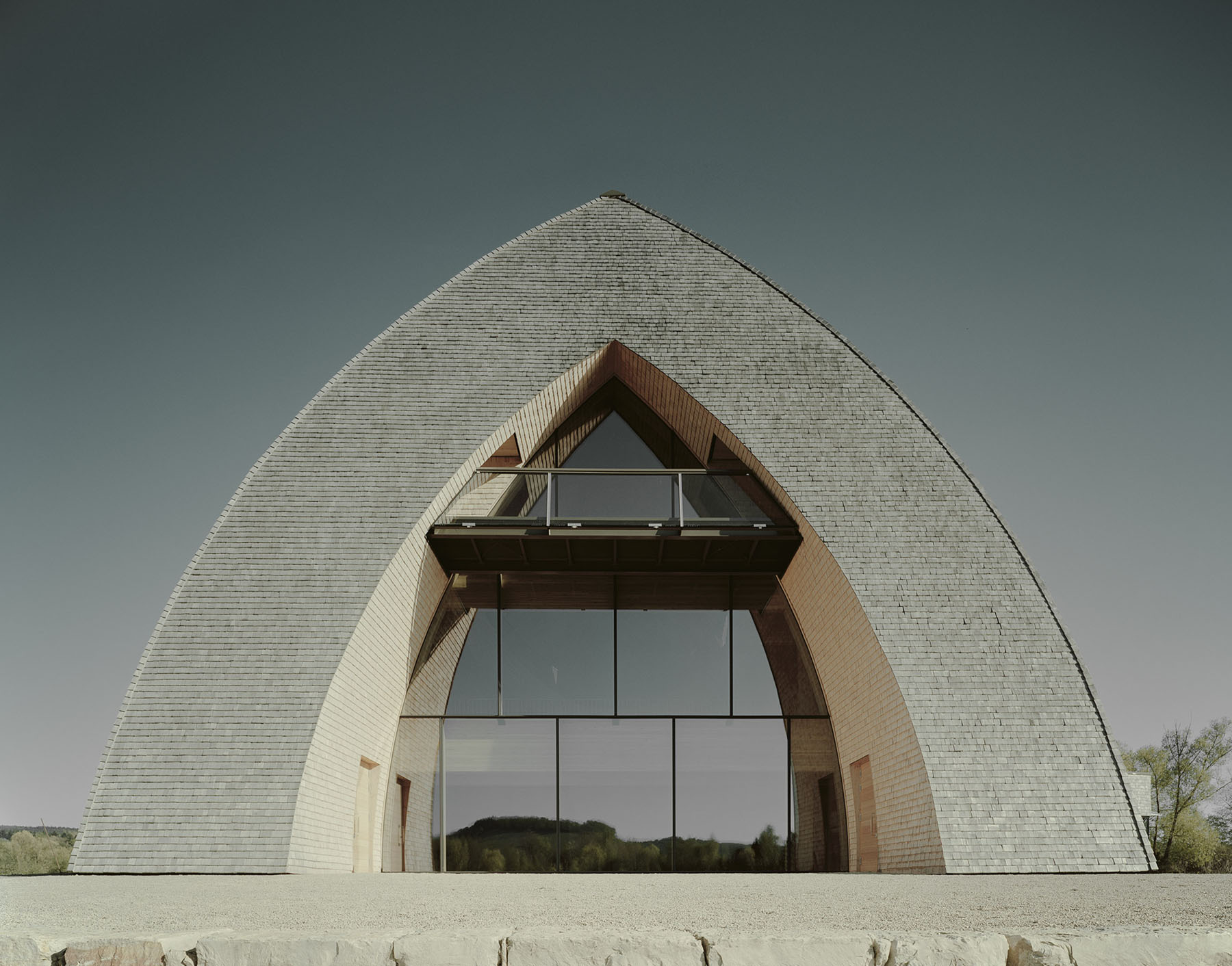 Biodiversum Nature Reserve Haff Réimech by Valentiny HVP Architects — The Biodiversum's monolithic arched facade presents a symmetrical composition dominated by a massive pitched-roof form clad in horizontal slate-grey tile...
