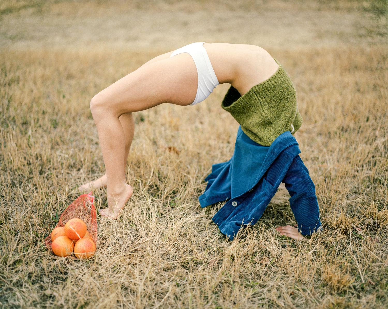 Infinite Tenderness by Peyton Fulford — A figure in a green knit sweater and blue denim jacket bends forward in an inverted pose across dry grass, head obscured by fabric.
