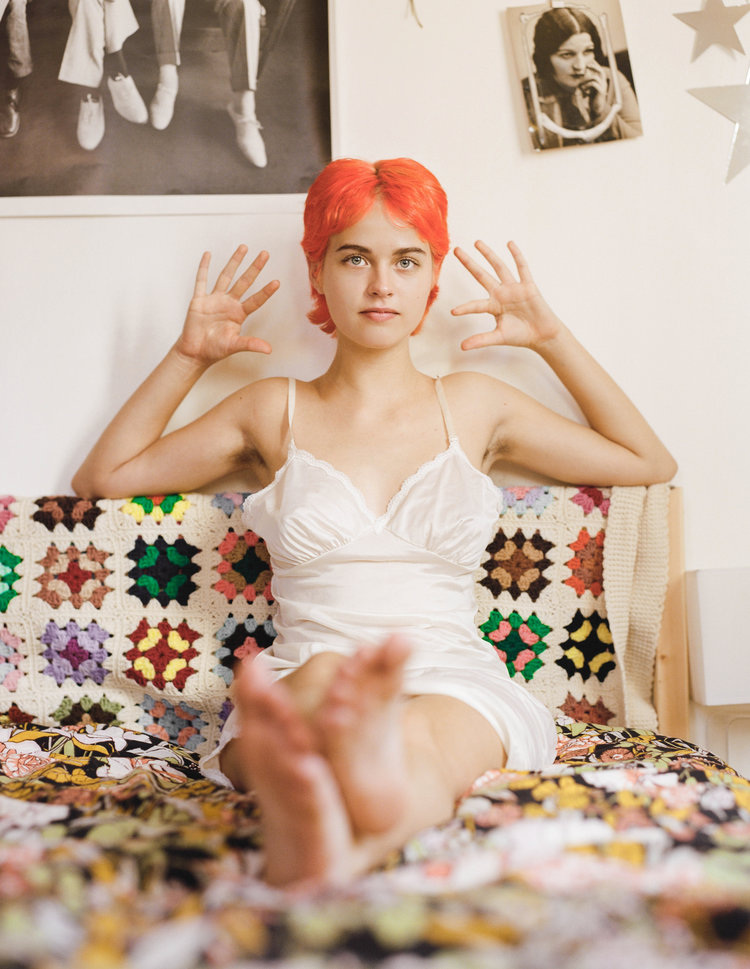 Infinite Tenderness by Peyton Fulford — A portrait photograph centered on a subject with vivid orange-red hair wearing a white wrap dress, seated on a patterned textile surface.
