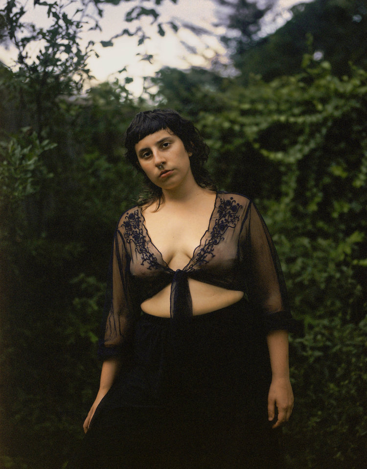 Infinite Tenderness by Peyton Fulford — A portrait photograph of a single figure positioned centrally against a densely vegetated background of dark green foliage.