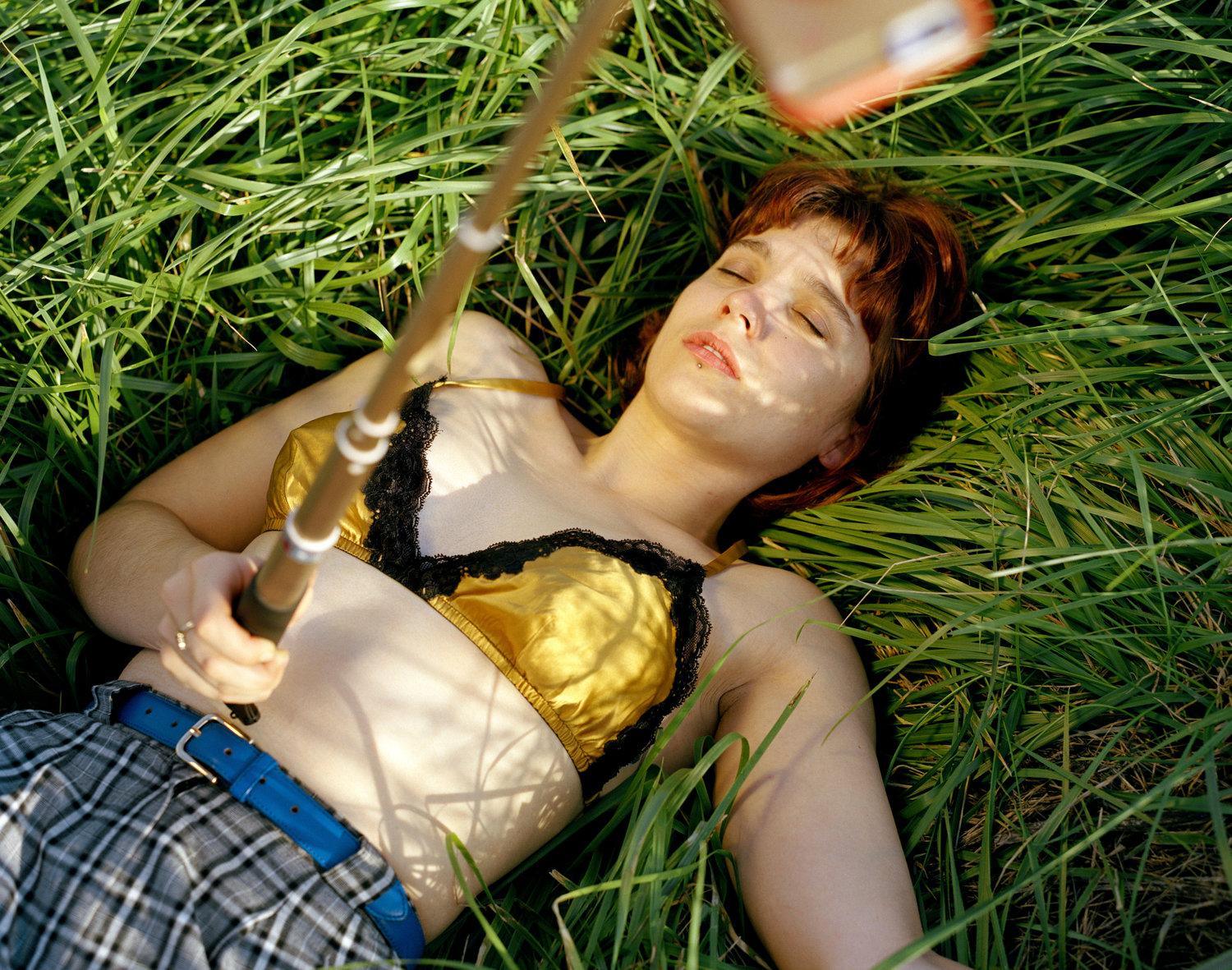 Infinite Tenderness by Peyton Fulford — A reclining figure with reddish-brown hair lies on grass wearing a gold satin bra with black lace trim and blue plaid fabric beneath.