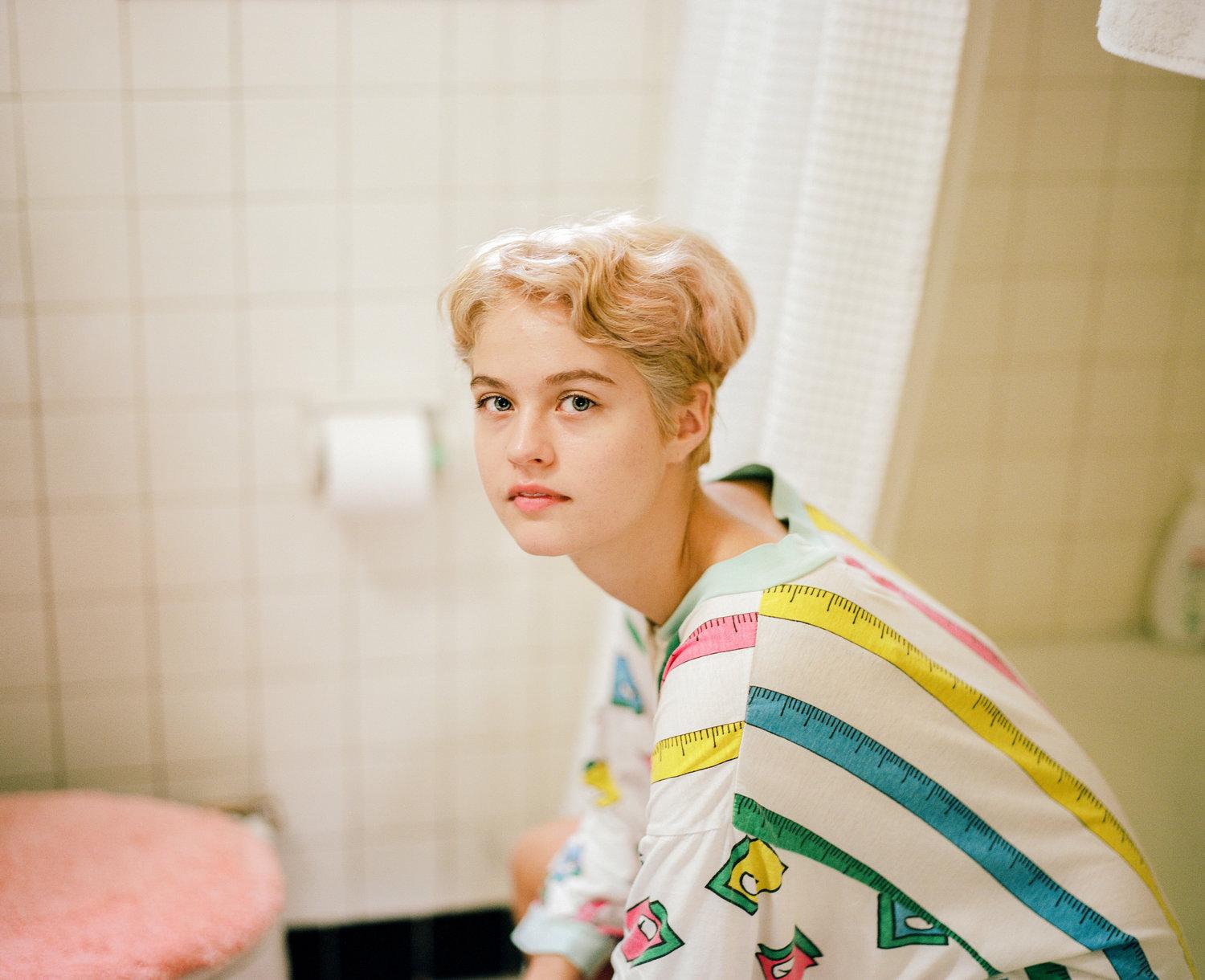 Infinite Tenderness by Peyton Fulford — A portrait photograph capturing a figure in a bathroom setting, wearing a cream-colored shirt printed with measurement tape motifs in yellow, pink, teal,...